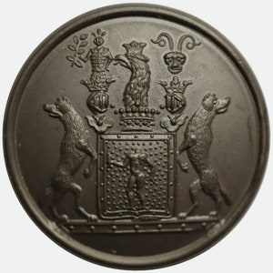 Könnte beinhalten: Ein dunkelbrauner Metallknopf mit einem erhabenen Design, das ein Wappen zeigt. Das Wappen enthält einen Schild mit einer Figur darin, zwei Hunde auf beiden Seiten und eine Krone über dem Schild.