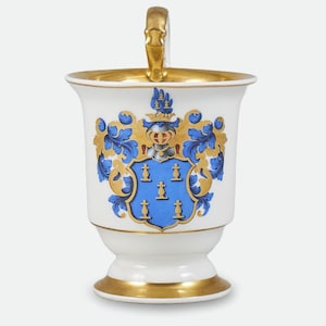 Könnte beinhalten: Eine weiße Porzellan Tasse mit goldenem Henkel und Verzierungen. Die Tasse zeigt ein blaues und goldenes Wappen mit einer Krone und floralen Akzenten.