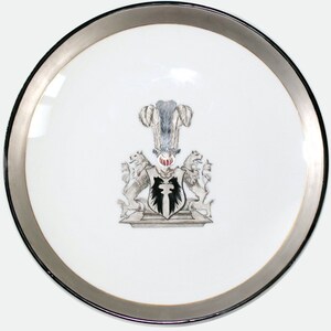 May include: A white porcelain plate with a silver rim features a black and white coat of arms with a crest. The crest is a black and white feathered helmet with a red and white face.