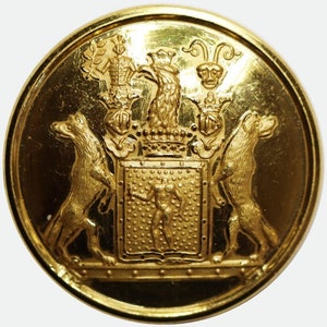 Könnte beinhalten: Ein goldfarbener, kreisförmiger Knopf mit einem geprägten Wappen-Design. Das zentrale Schild zeigt eine menschliche Figur, flankiert von zwei stilisierten Tieren. Der Rand und die Details des Knopfes sind stark reflektierend.