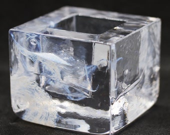 May include: A clear, square glass candle holder with a hollowed-out centre. The glass has a slightly swirled, textured appearance with white and blue accents. The candle holder is on a black surface.