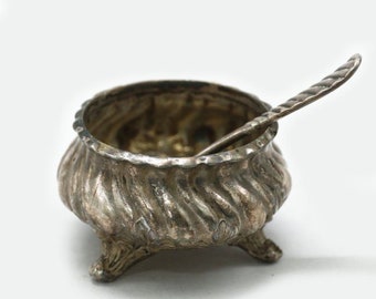 Puede incluir: Un salero de plata con una pequeña cuchara, que presenta un diseño decorativo en espiral y tres patas.