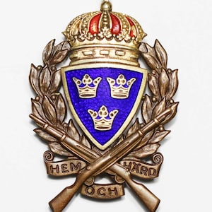 May include: A gold and bronze military badge with a blue shield featuring three gold crowns. The shield is surrounded by a wreath of leaves and crossed rifles. The text "HEM OCH HÅRD" is inscribed on a banner below the rifles.