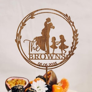 Może przedstawiać: Drewniany topper na tort z sylwetką rodziny czteroosobowej, pary i dwojga dzieci, z tekstem "The Browns 05.05.2025" wokół koła. Topper znajduje się na białym torcie ozdobionym owocami i kwiatami.