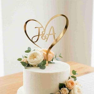 Może przedstawiać: Dwupiętrowy biały tort weselny ozdobiony brzoskwiniowymi i białymi różami oraz zielenią. Złoty topper w kształcie serca z inicjałami "J & A" znajduje się na wierzchu ciasta, dodając elegancji temu uroczystemu deserowi.