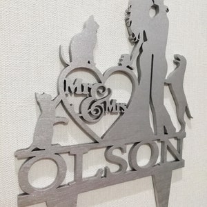 May include: Silver wedding cake topper featuring a couple, two cats, and a dog. The couple are holding hands and the dog is standing on its hind legs. The topper has a heart with the words "Mr & Mrs Olson" on it.