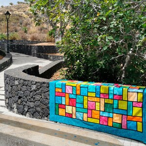 Pu&ograve; includere: Una trapunta colorata a patchwork con un design geometrico in tonalit&agrave; di blu, verde, giallo, arancione, rosa e rosso. La trapunta &egrave; fatta di quadrati e rettangoli ed &egrave; appoggiata su un muro di pietra.