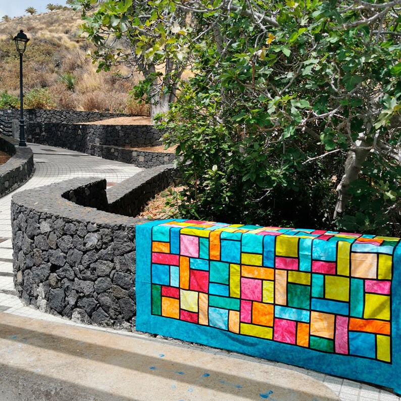 Pu&ograve; includere: Una trapunta colorata a patchwork con un design geometrico in tonalit&agrave; di blu, verde, giallo, arancione, rosa e rosso. La trapunta &egrave; fatta di quadrati e rettangoli ed &egrave; appoggiata su un muro di pietra.