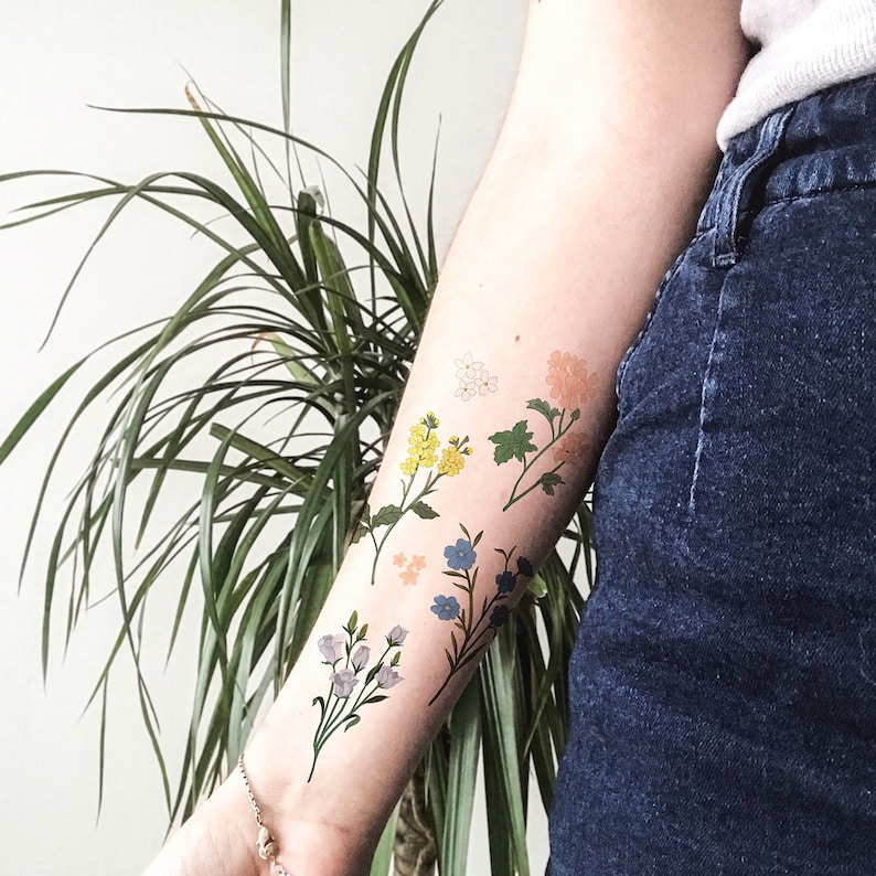 Pu&ograve; includere: Un braccio con un tatuaggio temporaneo di fiori colorati. I fiori sono in tonalit&agrave; di giallo, blu, rosa e arancio. Il tatuaggio &egrave; sull'avambraccio ed &egrave; visibile contro la pelle della persona.