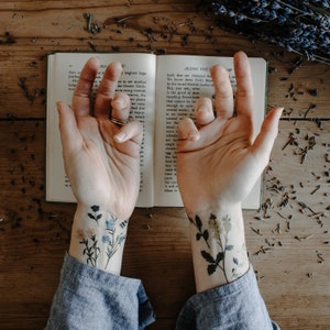 Pu&ograve; includere: Gli avambracci di una persona con due tatuaggi temporanei di delicati motivi floreali. I tatuaggi sono sui polsi sinistro e destro. La persona tiene un libro aperto con del testo sulle  fiori. Il libro &egrave; su una superficie di legno con fiori di lavanda secchi sparsi intorno.