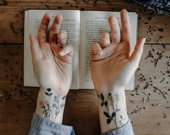 May include: A person's forearms with two temporary tattoos of delicate floral designs. The tattoos are on the left and right wrists. The person is holding an open book with text about flowers. The book is on a wooden surface with dried lavender flowers scattered around it.