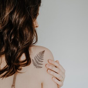 May include: A woman with long brown hair has a fern tattoo on her left shoulder.