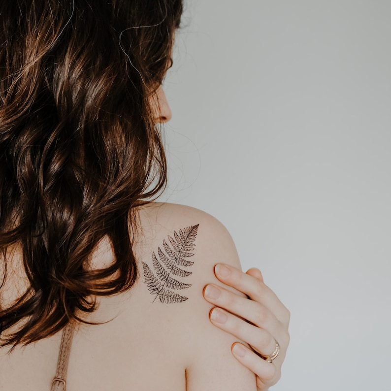 May include: A woman with long brown hair has a fern tattoo on her left shoulder.