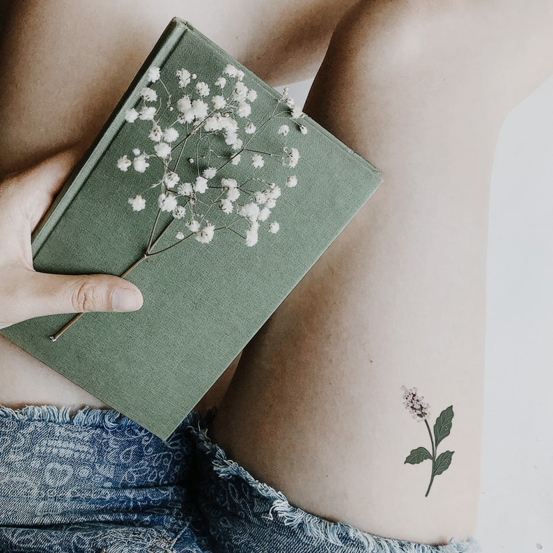Pu&ograve; includere: Una persona tiene in mano un libro verde con un rametto di fiori bianchi di gipsofila. Un piccolo tatuaggio di un fiore viola &egrave; sulla sua gamba.