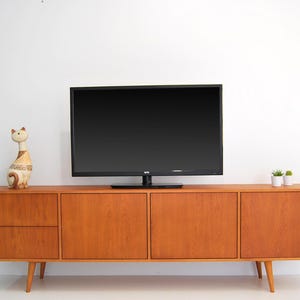 Puede incluir: Un mueble de TV de madera de estilo moderno de mediados de siglo con un televisor de pantalla plana negro. El mueble tiene cuatro puertas y dos cajones, con una figura decorativa de gato a la izquierda. Dos pequeñas plantas en macetas a la derecha.
