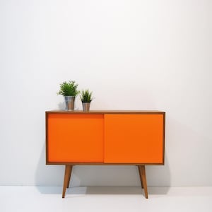 Könnte beinhalten: Ein Schrank im Stil der Mid-Century Modern mit zwei Schiebetüren. Der Schrank ist aus Holz mit einer natürlichen Oberfläche und hat orangefarbene Türen. Der Schrank steht auf vier Beinen und hat zwei Topfpflanzen oben drauf.
