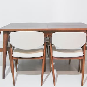 Könnte beinhalten: Ein Esstisch aus Holz mit einer braunen Tischplatte und vier weißen Stühlen. Der Tisch hat ein schlichtes Design mit geraden Beinen und einer rechteckigen Tischplatte. Die Stühle haben eine geschwungene Rückenlehne und einen gepolsterten Sitz.