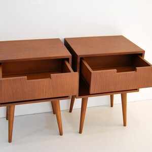 May include: Two wooden nightstands with a single drawer each. The drawer on the right nightstand is open, revealing the empty interior. The nightstands have a mid-century modern design with tapered legs.