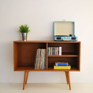 Puede incluir: Un mueble de madera para discos de estilo moderno de mediados de siglo con un tocadiscos en la parte superior. El mueble tiene dos estantes y está sobre cuatro patas. Los estantes están llenos de discos y libros.