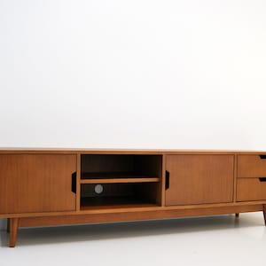 Puede incluir: Consola de medios de madera de estilo moderno de mediados de siglo. La consola tiene dos puertas, un estante abierto y dos cajones. La madera tiene un acabado c&aacute;lido y natural. La consola est&aacute; soportada por patas anguladas.