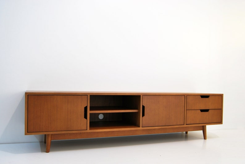 Puede incluir: Consola de medios de madera de estilo moderno de mediados de siglo. La consola tiene dos puertas, un estante abierto y dos cajones. La madera tiene un acabado c&aacute;lido y natural. La consola est&aacute; soportada por patas anguladas.