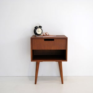 May include: A wooden nightstand with a single drawer and a lower shelf. The nightstand has four legs and is finished in a rich brown color. A black alarm clock and a pair of glasses are on top of the nightstand.