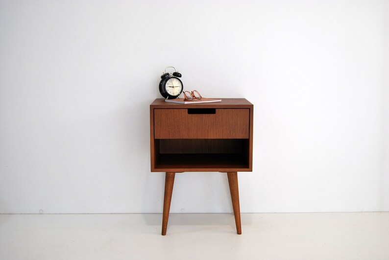 May include: A wooden nightstand with a single drawer and a lower shelf. The nightstand has four legs and is finished in a rich brown color. A black alarm clock and a pair of glasses are on top of the nightstand.