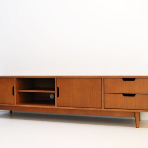 Puede incluir: Consola de medios de madera de estilo moderno de mediados de siglo con acabado marr&oacute;n claro. La consola tiene dos puertas, dos cajones y una zona de estanter&iacute;a abierta. Los cajones y las puertas tienen tiradores empotrados y la unidad se apoya en patas anguladas. Mide unos 180 cm de largo.