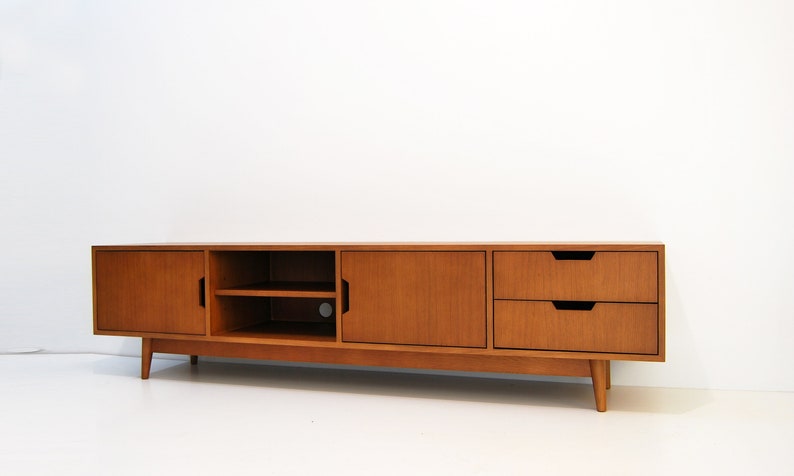 Puede incluir: Consola de medios de madera de estilo moderno de mediados de siglo con acabado marr&oacute;n claro. La consola tiene dos puertas, dos cajones y una zona de estanter&iacute;a abierta. Los cajones y las puertas tienen tiradores empotrados y la unidad se apoya en patas anguladas. Mide unos 180 cm de largo.