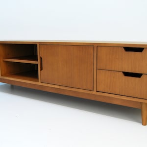 Puede incluir: Consola de medios de madera de estilo moderno de mediados de siglo, de color marr&oacute;n claro. Cuenta con dos cajones, dos puertas de armario y una balda abierta. La unidad se apoya en cuatro patas c&oacute;nicas. Mide unos 180 cm de largo.