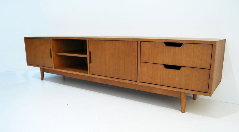 Puede incluir: Consola de medios de madera de estilo moderno de mediados de siglo, de color marr&oacute;n claro. Cuenta con dos cajones, dos puertas de armario y una balda abierta. La unidad se apoya en cuatro patas c&oacute;nicas. Mide unos 180 cm de largo.