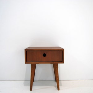 Puede incluir: Una mesita de noche de madera con un caj&oacute;n y cuatro patas. El caj&oacute;n tiene un peque&ntilde;o recorte redondo en la parte delantera.