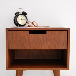 May include: A wooden bedside table with a single drawer and a lower shelf. The table has a dark brown finish and is sitting on four legs. A black alarm clock and a pair of glasses are sitting on top of the table.