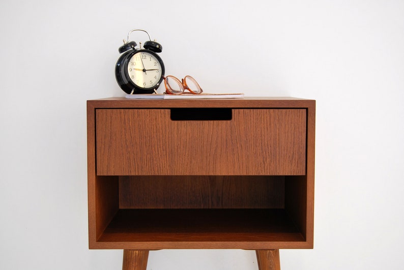 May include: A wooden bedside table with a single drawer and a lower shelf. The table has a dark brown finish and is sitting on four legs. A black alarm clock and a pair of glasses are sitting on top of the table.