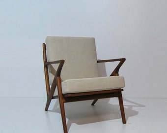 May include: A mid-century modern armchair with a light beige fabric upholstery and a dark wood frame. The chair has a unique angled design with a wide, comfortable seat.