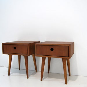 Puede incluir: Dos mesitas de noche de madera con un caj&oacute;n y patas c&oacute;nicas. Las mesitas de noche son de un color marr&oacute;n intenso y tienen un dise&ntilde;o sencillo y moderno de mediados de siglo.