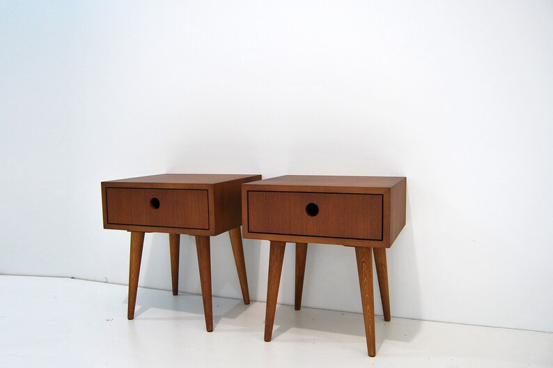 Puede incluir: Dos mesitas de noche de madera con un caj&oacute;n y patas c&oacute;nicas. Las mesitas de noche son de un color marr&oacute;n intenso y tienen un dise&ntilde;o sencillo y moderno de mediados de siglo.