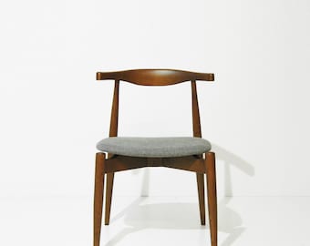 Puede incluir: Una silla de madera de estilo moderno de mediados de siglo con un asiento tapizado gris. La silla tiene un respaldo curvo y cuatro patas anguladas. La madera tiene un acabado cálido y natural, y el asiento es gris.