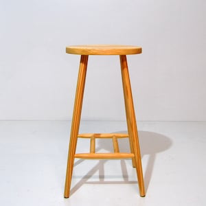 May include: A simple wooden stool with a round seat and four legs. The stool is made of natural wood and has a light brown color.