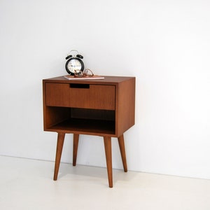 May include: A mid-century modern style wooden nightstand with a single drawer and a lower shelf. The nightstand has four tapered legs and is finished in a rich brown color. A black alarm clock and a pair of glasses are on top of the nightstand.