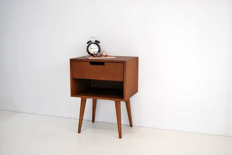May include: A mid-century modern style wooden nightstand with a single drawer and a lower shelf. The nightstand has four tapered legs and is finished in a rich brown color. A black alarm clock and a pair of glasses are on top of the nightstand.
