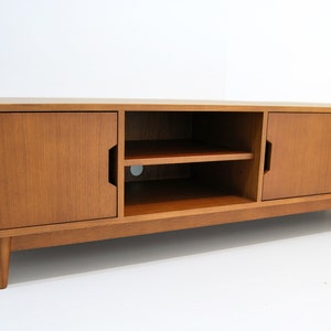 Puede incluir: Consola de medios de madera de estilo moderno de mediados de siglo. La consola tiene dos puertas, un estante abierto y dos cajones. La madera tiene un acabado marr&oacute;n claro y las patas son c&oacute;nicas. La consola est&aacute; dise&ntilde;ada para una sala de estar.