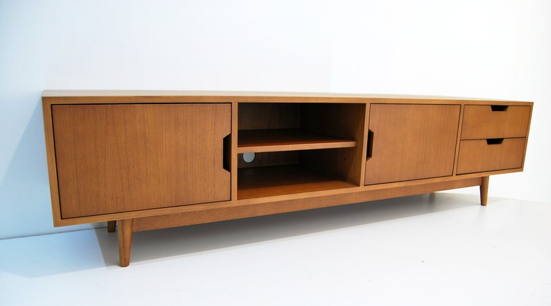 Puede incluir: Consola de medios de madera de estilo moderno de mediados de siglo. La consola tiene dos puertas, un estante abierto y dos cajones. La madera tiene un acabado marr&oacute;n claro y las patas son c&oacute;nicas. La consola est&aacute; dise&ntilde;ada para una sala de estar.