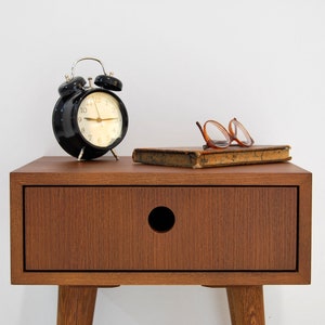 Puede incluir: Una mesita de noche de madera con un caj&oacute;n y una perilla redonda. Un despertador negro con detalles plateados se encuentra en la parte superior de la mesita de noche, junto con un libro y un par de gafas.