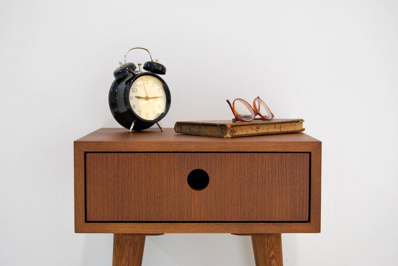 Puede incluir: Una mesita de noche de madera con un caj&oacute;n y una perilla redonda. Un despertador negro con detalles plateados se encuentra en la parte superior de la mesita de noche, junto con un libro y un par de gafas.