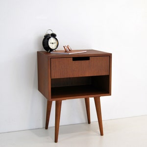 May include: A wooden bedside table with a single drawer and a lower shelf. The table has four tapered legs and is finished in a rich brown color. A black alarm clock and a pair of glasses are on top of the table.