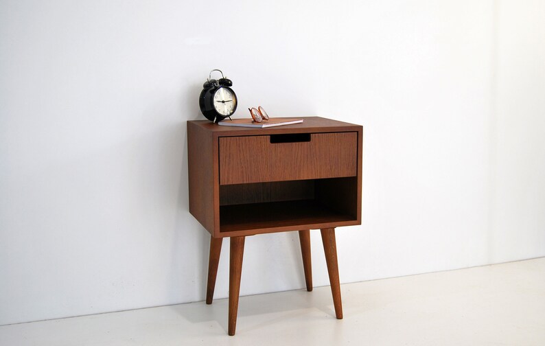 May include: A wooden bedside table with a single drawer and a lower shelf. The table has four tapered legs and is finished in a rich brown color. A black alarm clock and a pair of glasses are on top of the table.
