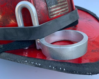 Op de afbeelding: Een zilveren metalen drinkhouder bevestigd aan een rode brandweerhelm met een zwarte band. De helm heeft een vervaagde Amerikaanse vlag sticker en gele stickers met tekst.