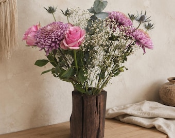 May include: A bouquet of pink roses, purple chrysanthemums, blue sea holly, baby's breath, and eucalyptus leaves arranged in a dark brown, cylindrical wooden vase with a textured, bark-like exterior surface and vertical grooves.