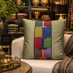 May include: A square olive green throw pillow featuring a central vertical panel of multi-coloured fabric patches in shades of red, blue, green, purple, brown, and orange, separated by thin black stitching and bordered by black piping.